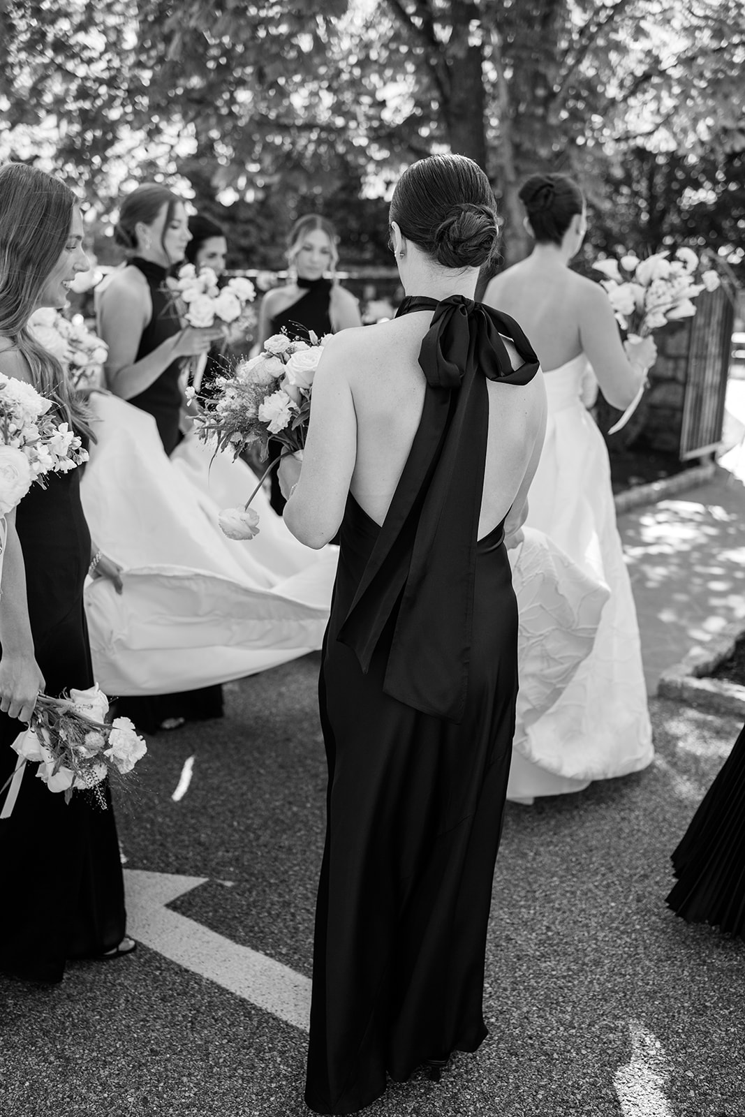Black and white film photo of bridesmaids carrying the bride's gown train as they walk outdoors.